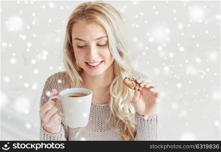 morning, leisure and people concept - happy young woman with cup of tea eating cookie in bed at home bedroom over snow. happy woman with tea and cookies in bed at home