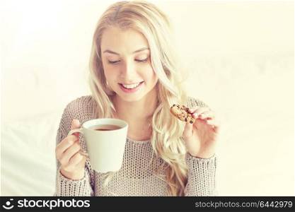 morning, leisure and people concept - happy young woman with cup of tea eating cookie in bed at home bedroom. happy woman with tea and cookies in bed at home