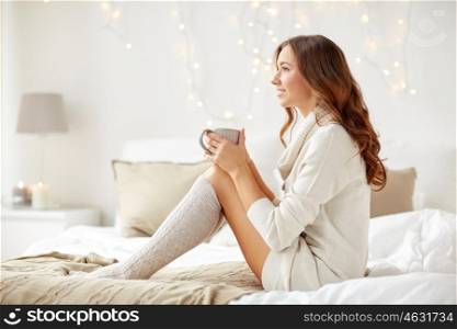 morning, leisure, christmas, winter and people concept - happy young woman with cup of coffee or tea in bed at home bedroom