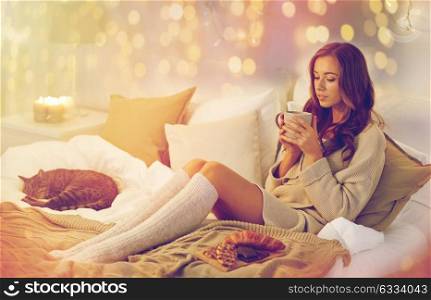 morning, leisure, christmas, winter and people concept - happy young woman with cup of coffee or tea and food in bed at home bedroom. happy woman with cup of coffee in bed at home