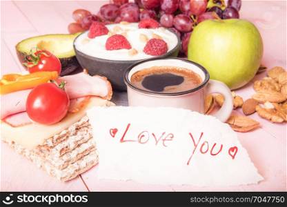 Morning meal with healthy food, fruits, a bowl of yogurt with cereal, vegetables, whole-wheat crispbread, nuts, a cup of coffee and a love you message.