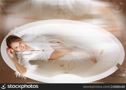 Morning of the bride. Happy beautiful young woman is wearing in a white long veil, robe and underwear sitting laughing and having fun in the bath on a white background, relax. wedding day.. Morning of the bride. Happy beautiful young woman is wearing in a white long veil, robe and underwear sitting laughing and having fun in the bath on a white background, relax. wedding day