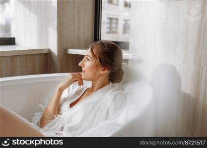 Morning of the bride. Happy beautiful young woman is wearing in a white long veil, robe and underwear sitting laughing and having fun in the bath on a white background, relax. wedding day.. Morning of the bride. Happy beautiful young woman is wearing in a white long veil, robe and underwear sitting laughing and having fun in the bath on a white background, relax. wedding day