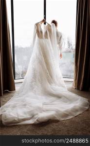Morning of the bride. the bride is holding a luxury white long dress in her hands by the large window in hotel room. back view. Morning of the bride. the bride is holding a luxury white long dress in her hands by the large window in hotel room. back view.