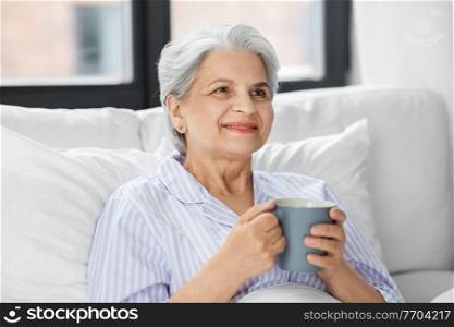 morning, old age and people concept - happy smiling senior woman in pajamas with cup of coffee sitting on bed at home bedroom. old woman with cup of coffee in bed at home