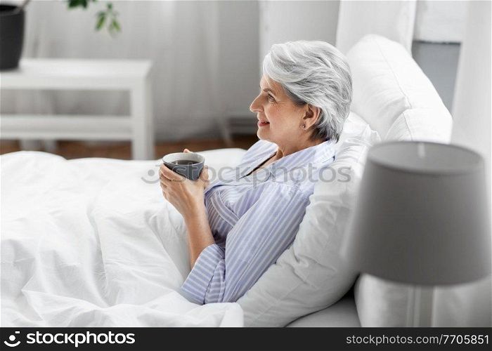 morning, old age and people concept - happy smiling senior woman in pajamas with cup of coffee sitting on bed at home bedroom. old woman with cup of coffee in bed at home