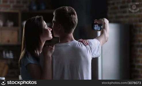 Morning photos in the kitchen. Rear view of loving couple embracing, kissing with tenderness while taking selfie photo on mobile phone. Young affectionate couple making self-portrait with phone at home.