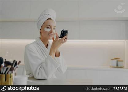 Morning skin care routine. Young beautiful woman in bathrobe and hair wrapped in towel applying cosmetic patches under eyes from dark circles and looking in compact mirror while standing in kitchen. Happy woman in white bathrobe with towel on head taking care of her skin after morning shower