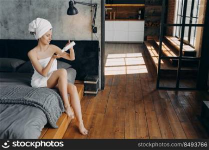 Morning skincare beauty ritual. Beautiful european woman wrapped in towel is sitting in bed. Slim girl applying body moisturizing lotion from bottle. Modern interior of bedroom. Bodycare and wellness.. Morning skincare beauty ritual. Beautiful european woman applying body moisturizing lotion.