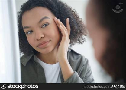 Morning skincare. Confident afro girl is applying cream to her face and looking into mirror. Skin cleansing and moisturizing with lotion. Daily beauty routine and femininity of teenage girl.. Morning skincare. Confident afro girl is applying cream to her face and looking into mirror.