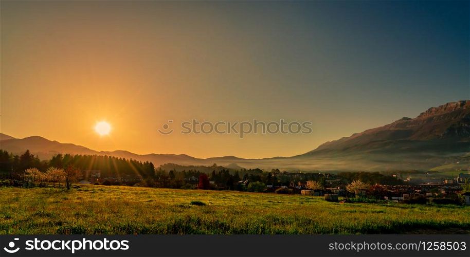 Morning sunrise over the mountain with clear blue sky. Grass field and pine forest in rural village near rock mountain. The fog covered the mountain in the morning. City in valley. Landscape meadow
