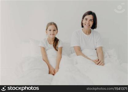 Morning time and awakening concept. Happy smiling mother and daughter sit in comfortable bed, dressed in white t shirts, awake with positive mood, glad to start new day, spend free time in bedroom.