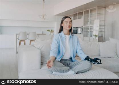 Morning yoga of disabled woman. Lady sitting in lotus pose on couch with her eyes closed. Attractive european girl has myoelectric cyber prosthesis. Modern artificial limb with nervous sensor control.. Morning yoga of disabled woman. Attractive european girl has myoelectric cyber prosthesis.