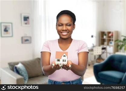 mortgage, real estate and accommodation concept - happy smiling african american woman holding house model over home room background. smiling african american woman holding house model