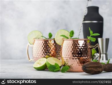 Moscow mule cocktail in a copper mug with lime and mint and wooden squeezer on light kitchen table background with black shaker and jigger.Space for text