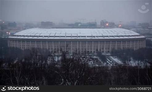 Moscow winter cityscape: Luzhniki Stadium with cars passing by near and bare trees in the foreground