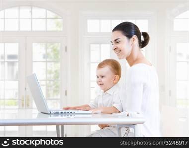 Mother and 2 years old baby boy sitting at the desk and using laptop computer. Indoor, brightly lit.