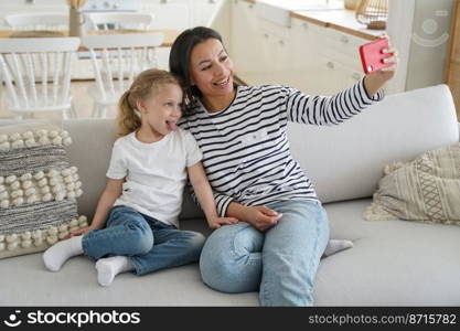 Mother and adopted little daughter, showing tongue, taking family pictures, looking at mobile phone camera, sitting on sofa. Happy mom having fun with preschool kid girl, takes selfie together at home. Mother, adopted little daughter, showing tongue, take family selfie together, using phone on couch