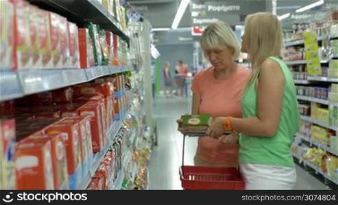 Mother and adult daughter buying food in the hypermarket. Women taking a package, checking it and putting into shopping basket