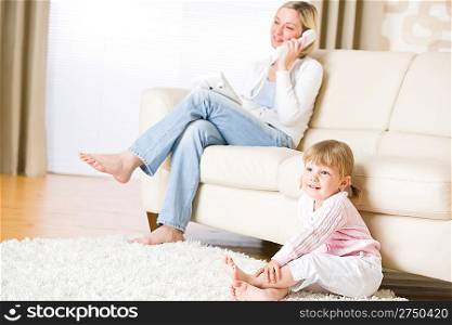 Mother and child in living room watch television, mother on phone in background on sofa