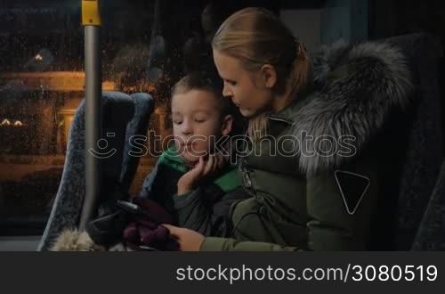 Mother and child passing the time with mobile during evening bus travel in winter. They watching movie or cartoons on cell