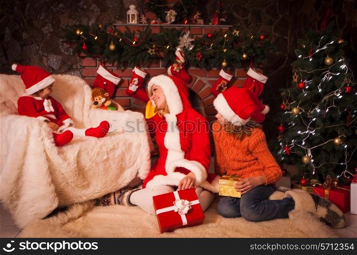 Mother and children are sitting near fireplace and christmas tree with gift boxes.