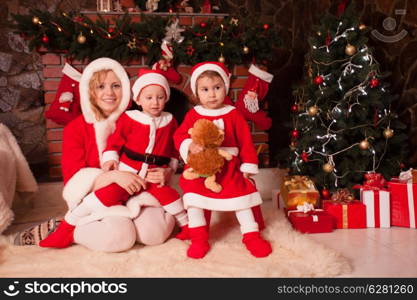 Mother and children are sitting near fireplace and christmas tree with gift boxes. Brother and sister in santa suits