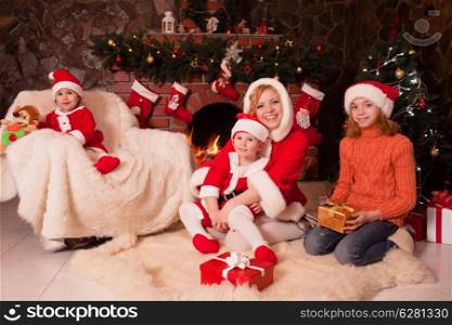 Mother and children are sitting near fireplace and christmas tree with gift boxes. Brother and sister in santa suits