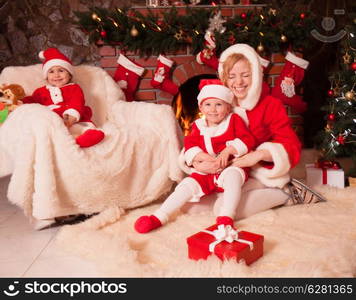 Mother and children are sitting near fireplace and christmas tree with gift boxes. Brother and sister in santa suits