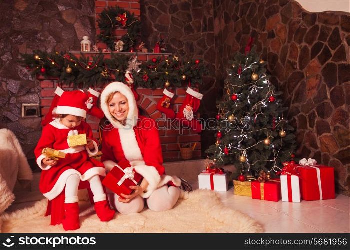 Mother and daughter are sitting near fireplace and christmas tree with gift boxes.