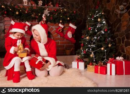 Mother and daughter are sitting near fireplace and christmas tree with gift boxes.