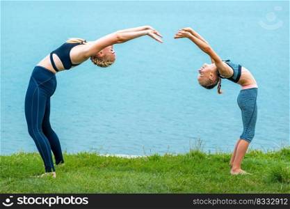 Mother and daughter doing gym exercises on the grass at the shore of the lake. Mother and daughter doing exercises