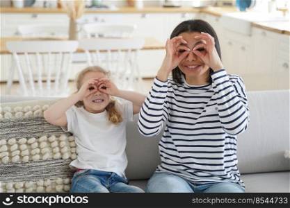 Mother and daughter fooling around do funny faces make binoculars with fingers, sitting on couch at home. Mum, little kid girl having fun together, showing glasses by hands, posing, looking at camera.. Mother, daughter fool around do funny faces make binoculars with fingers having fun together at home
