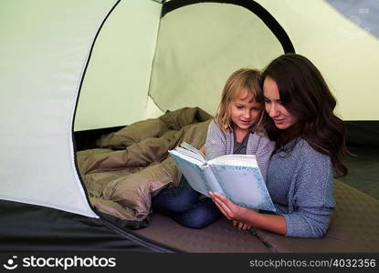 Mother and daughter in sleeping bag in tent, reading book together