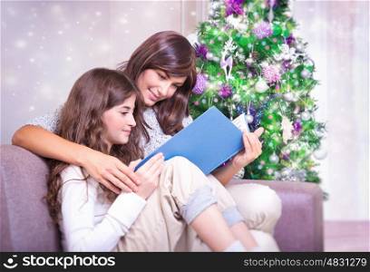 Mother and daughter sitting near Christmas tree and reading book, enjoying romantic fairy tale, happy family evening at home
