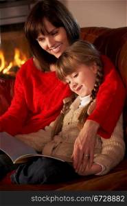 Mother And Daughter Sitting On Sofa And Reading Book By Cosy Log Fire