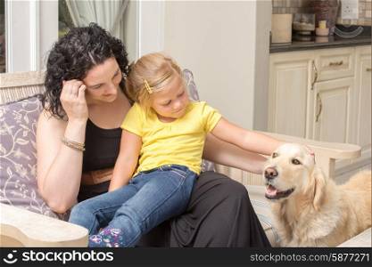 Mother and daughter sitting together on a chair on the porch of their home while stoking the golden retriever pet dog.