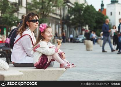 Mother and daughter spending time together, sitting in the center of town, little smiling girl holding and eating ice cream. Candid people, real moments, authentic situations
