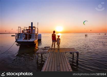 Mother and daughter standing on the pier in the background of the sea, ship and sunset. Ukrainian landscape