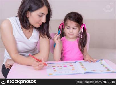 Mother and daughter study activity book with pencils on paper while sitting at pink table.Preschool and kindergarten education at home.Selective focus and small depth of field.. Mother and daughter study activity book with pencils on paper