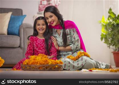 Mother and daughter together posing in front of camera while making flower garland on occasion