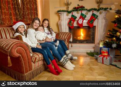 Mother and daughters sitting on sofa by the burning fireplace at house decorated for Christmas