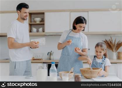Mother and father give eggs to daughter who prepares dough, busy cooking together during weekend, have happy moods, prepare food. Three family members at home. Parenthood and togetherness concept