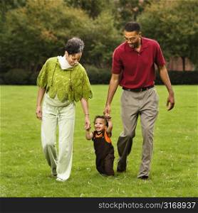 Mother and father helping toddler walk holding his hands in park.