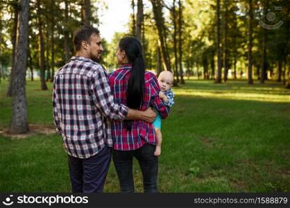 Mother and father walks together with little baby in summer park. Mom and dad with male kid on lawn, parents on picnic with child in the forest, family happiness. Mother and father walks together with little baby