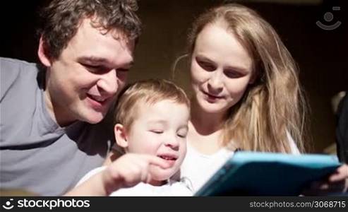 Mother and father watching boy playing game on touchpad. Happy and friendly family
