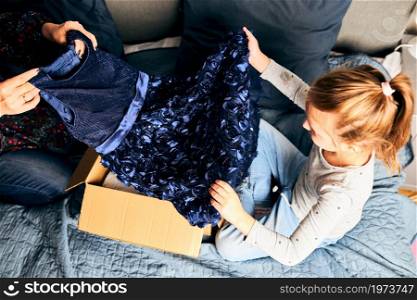 Mother and her daughter opening box with ordered dress at home on couch. Mom online shopper customer holding dress. Delivery service fashion concept