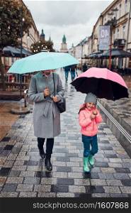 Mother and her little daughter holding the pink and blue umbrellas walking in a downtown on rainy gloomy autumn day. Woman and little girl holding the umbrellas walking in a downtown on rainy gloomy autumn day