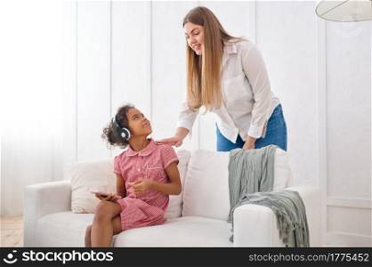 Mother and her little daughter in headphones on sofa in living room.Mom and female child leisures in their house together, good relationship, parental care. Mother and her little daughter in headphones
