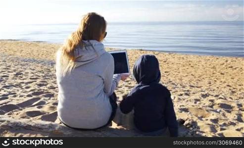 Mother and her little son on the beach. Boy touching the screen of tablet PC held by mother
